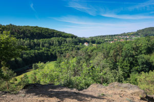Po obou stranách prochází zalesněné svahy podél Berounky. V popředí skalní plošina tvořící vyhlídku Kozí Bouda. V pozadí zástavba Bukovce.