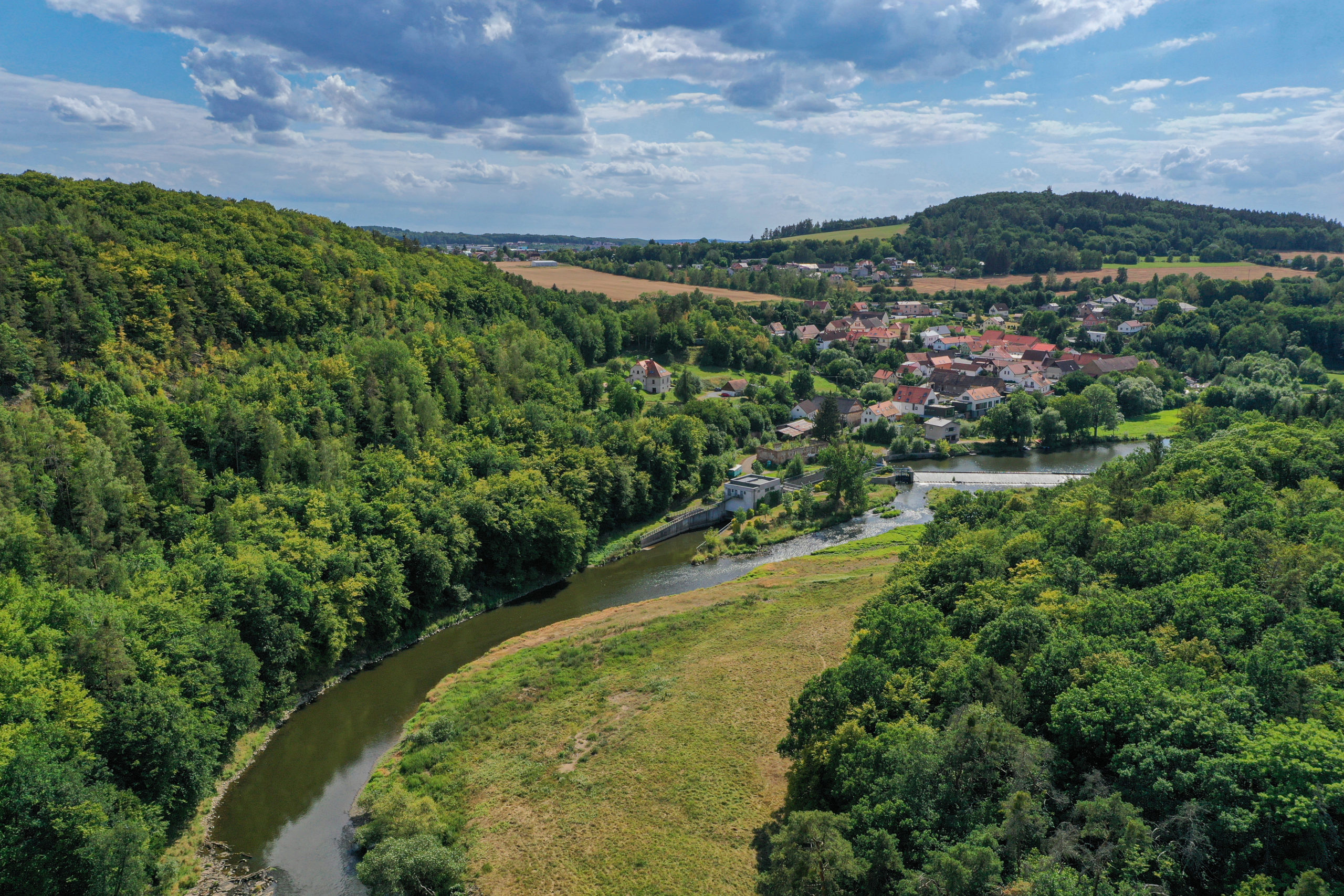 Pohled na část Zábělé a Bukovec z ptačí perspektivy z levého břehu Berounky, která šikmo protíná střední část snímku. Po obou stranách vidíme svahy s listnatými lesy, uprostřed rovinatá louka u řeky.