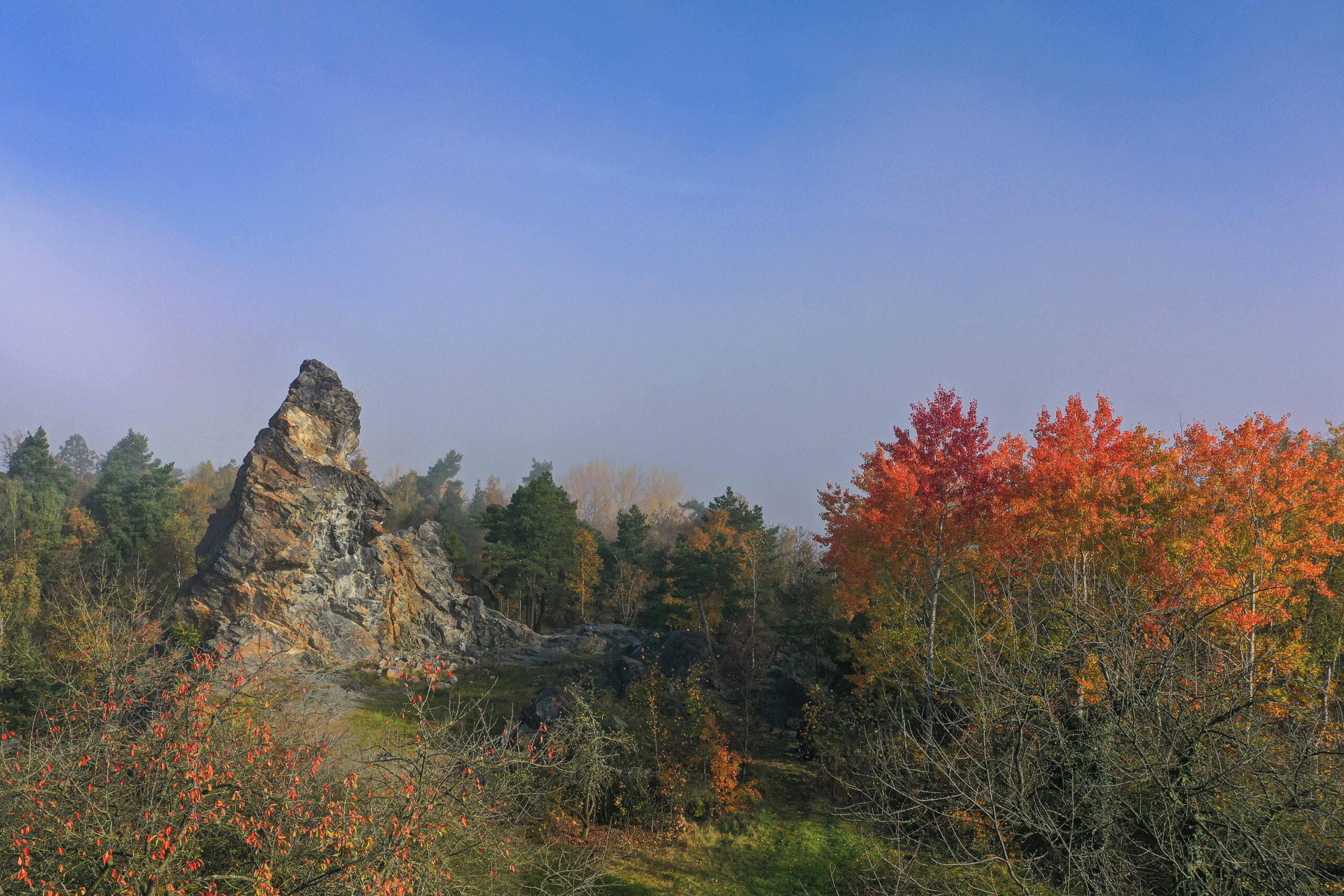 Pohled z ptačí perspektivy. V levé části se tyčí jehlancovitý skalní útvar, pravé části dominuje několik oranžově až červeně zbarvených listnatých stromů (osiky). V horní polovině snímku vidíme nebe s rozpouštějící se mlhou.