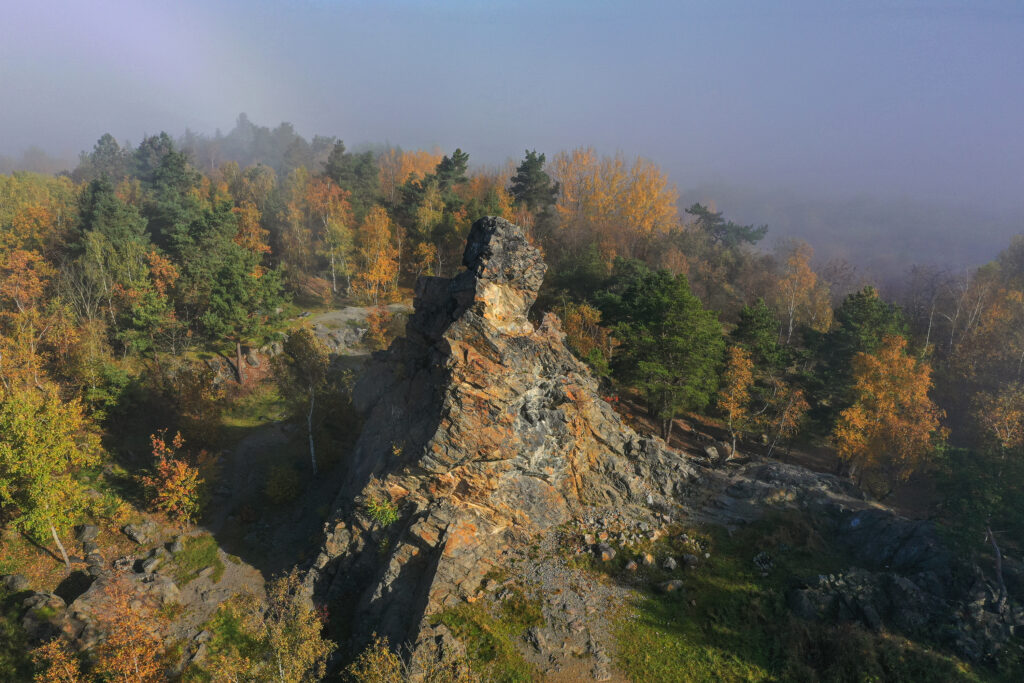 Pohled z ptačí perspektivy na jehlancovitý skalní útvar, u jehož paty navazují živě zelené plochy bývalého lomu. Vše obklopují porosty borovic s listnatými dřevinami v podzimních barvách.