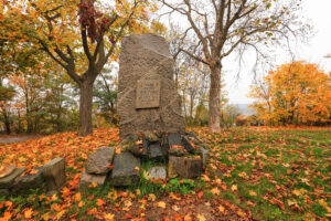Ve středu snímku stojící kamenný pomník s vystupující deskou. Okolo kosený trávník pokrytý zářivě barevnými listy. Zadní polovinu tvoří stromy v podzimních barvách.