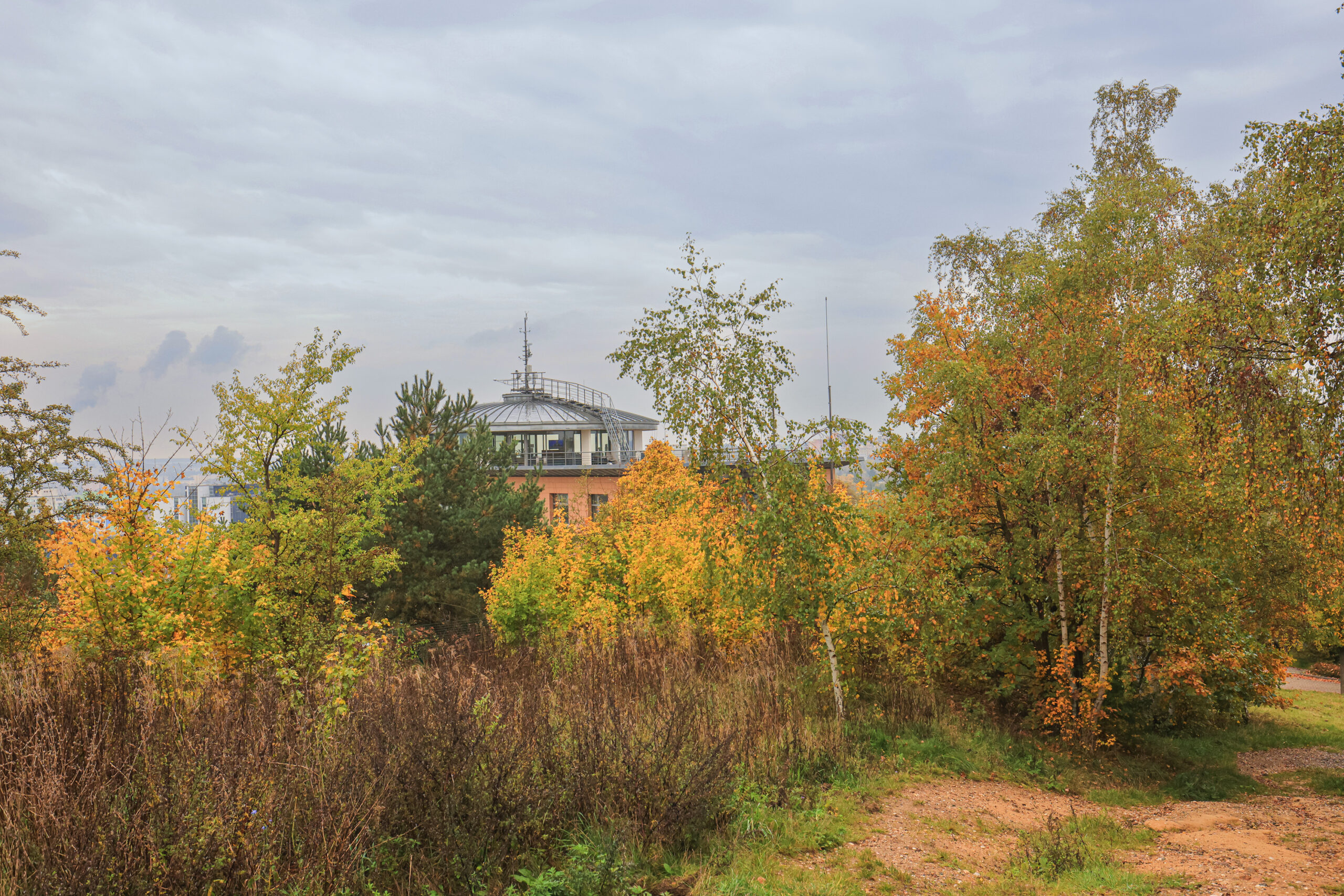 <h1>Vrch Mikulka – meteorologická stanice</h1><br />
V popředí rozvolněný porost náletových dřevin v podzimních barvách, za ním je nalevo od středu snímku viditelná okrouhlá, nízká věž meteorologické stanice. Pozadí tvoří zatažené nebe.